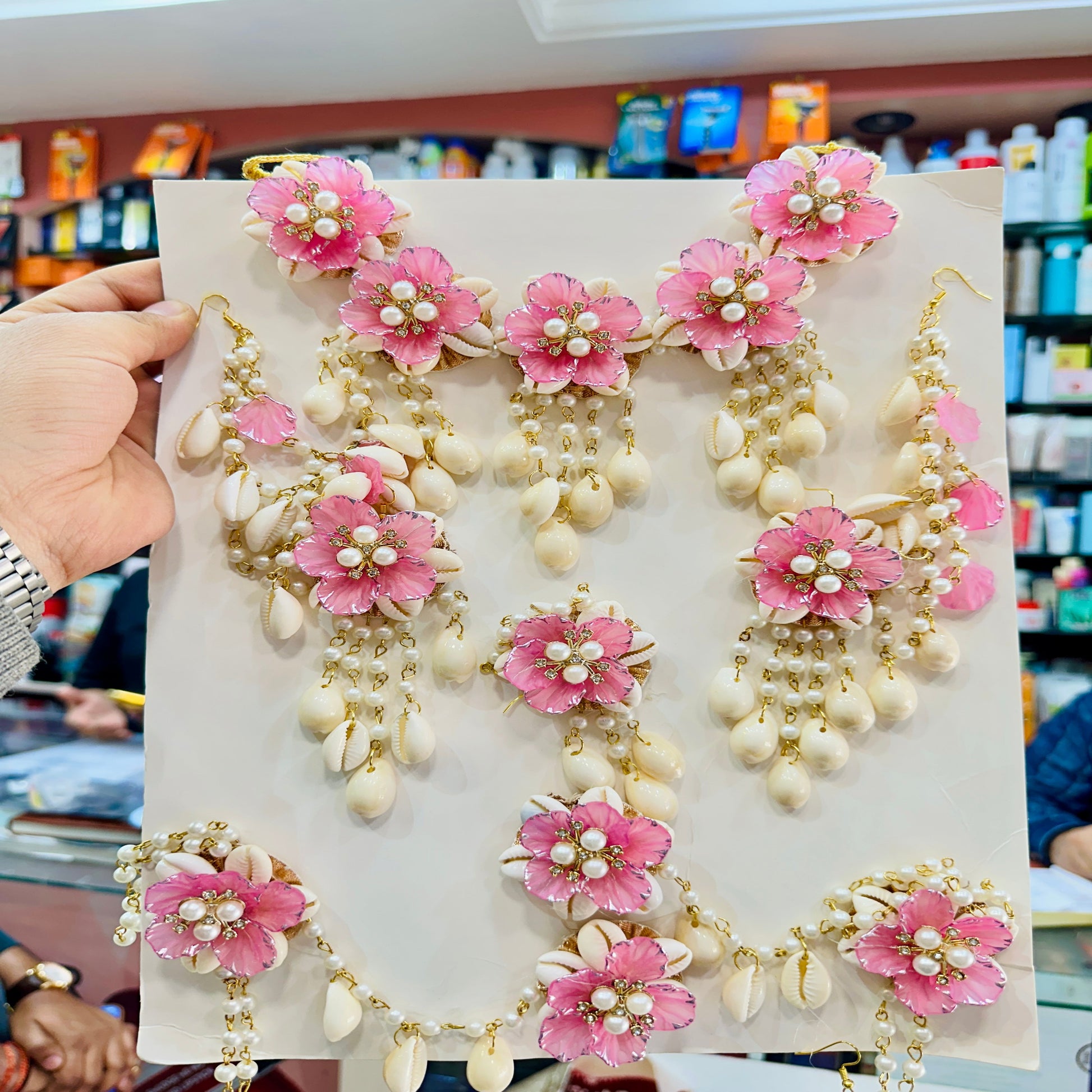 Decorative necklace with pink floral and pearl details on a white card, held in a store setting.